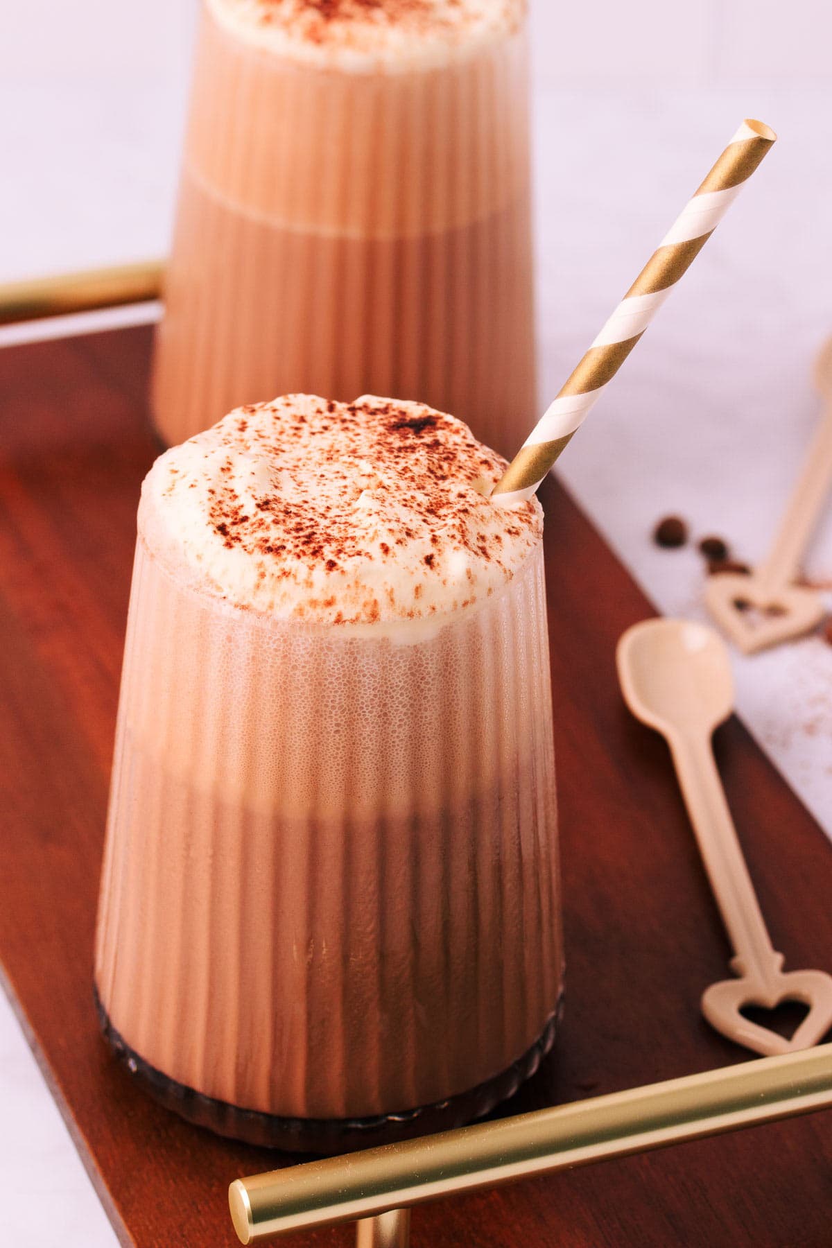 A mocha milkshake in a tapered glass with a ribbed texture, topped with whipped cream and a sprinkle of cocoa, with a gold and white striped paper straw on a dark wooden board with gold handles and a mocha-coloured ceramic spoon.