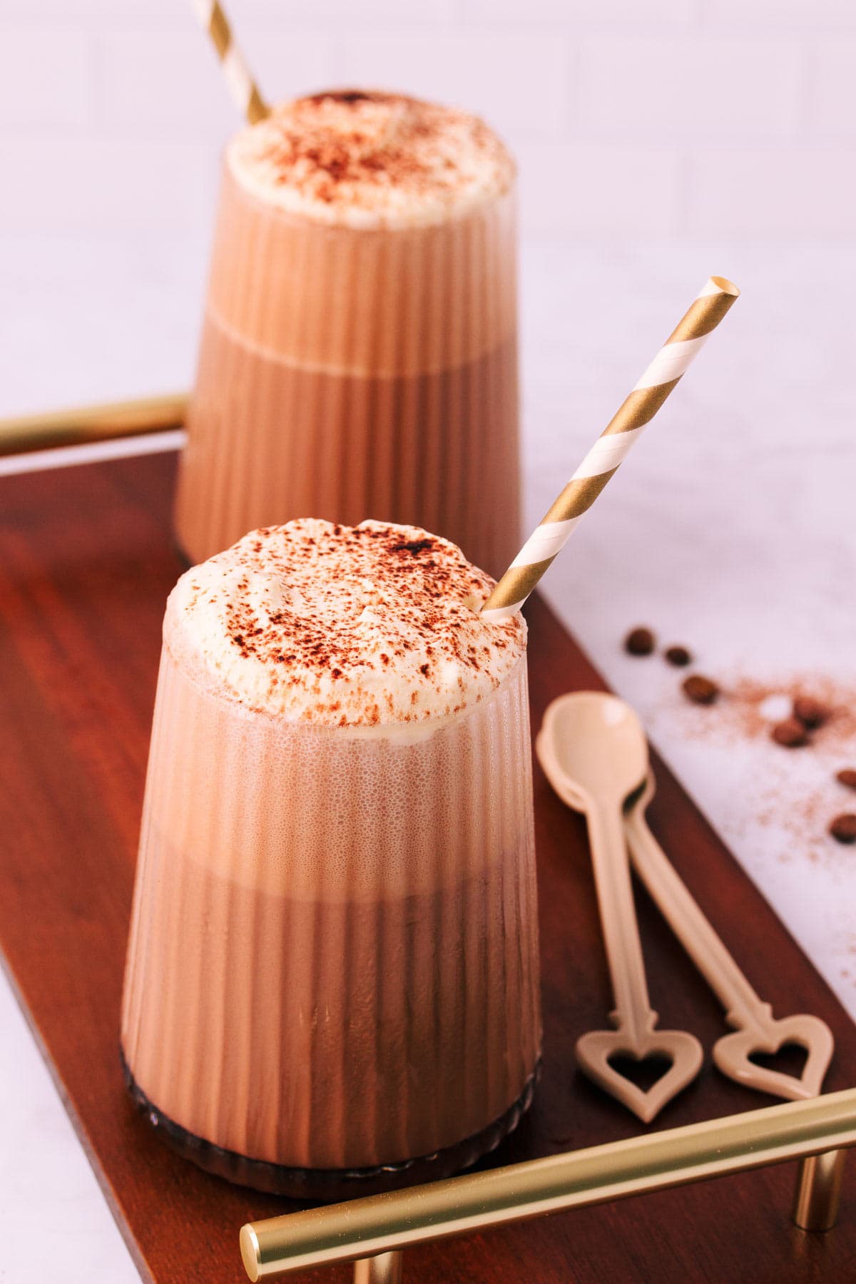 Two mocha milkshakes in glasses with a ribbed texture, topped with whipped cream and a sprinkle of cocoa, with gold and white striped paper straws on a dark wooden board with gold handles.