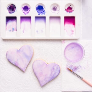Two heart shaped cookies covered with fondant icing and painted with pink and purple watercolour edible paint, with a paintbrush and paint palette on a grey marble background.