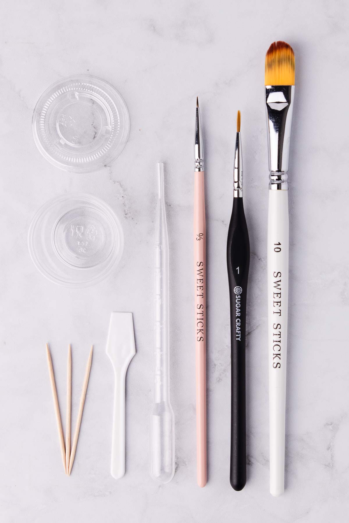 The tools and supplies used for making and using edible gold paint - paintbrushes, toothpicks, a clear plastic pipette and a small plastic container, on a light grey marble background.