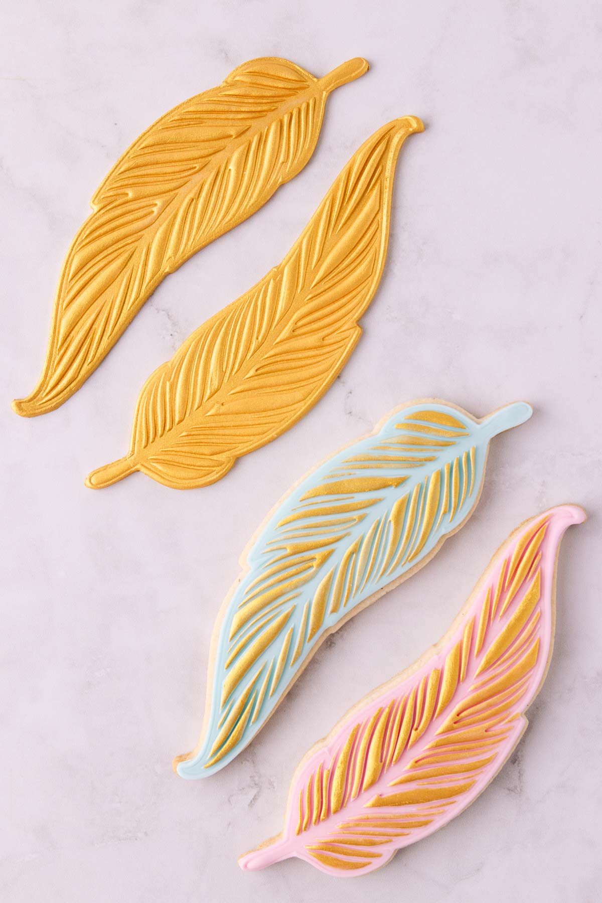 Two gold painted feather fondant shapes and two cookies, one with pink fondant and one with light blue fondant, both with gold painted details.