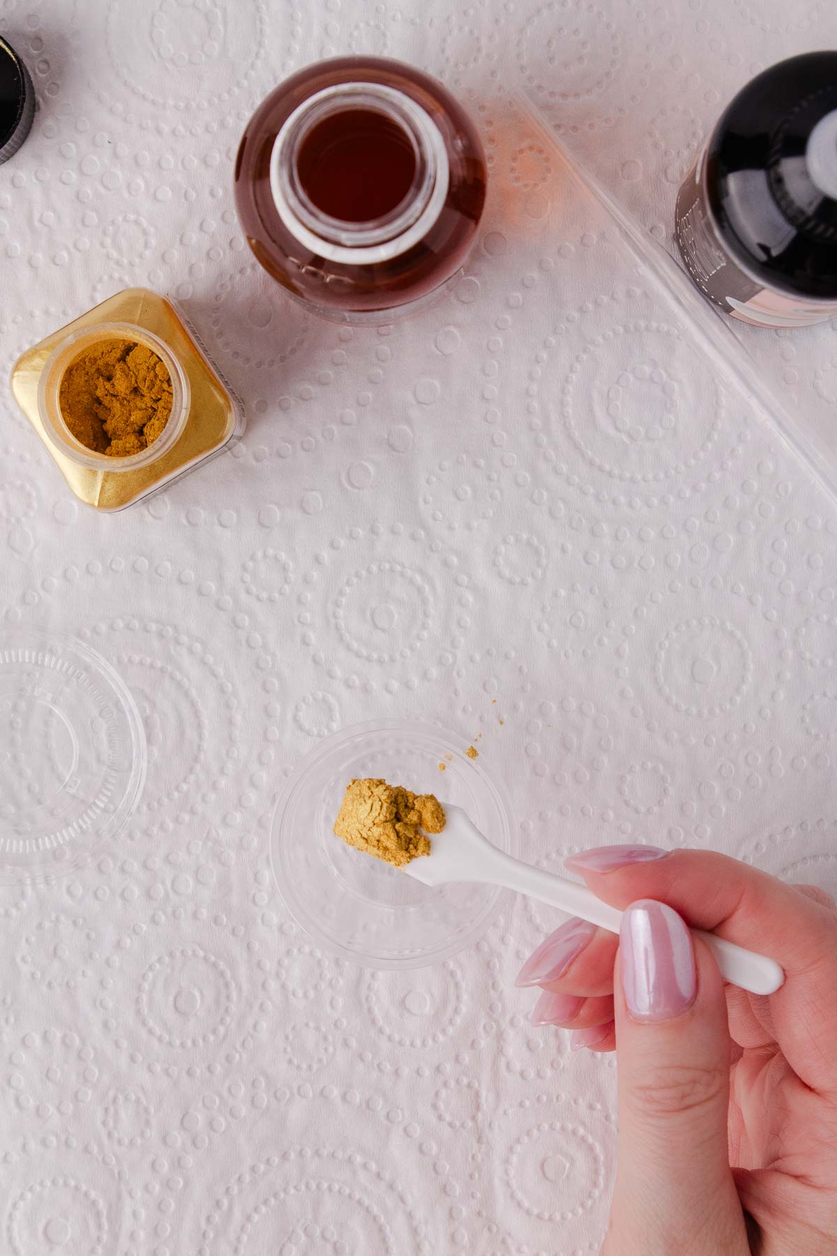 A hand holding a small white spoon with about 1/4 teaspoon of gold edible lustre powder on the end, over a small plastic container.