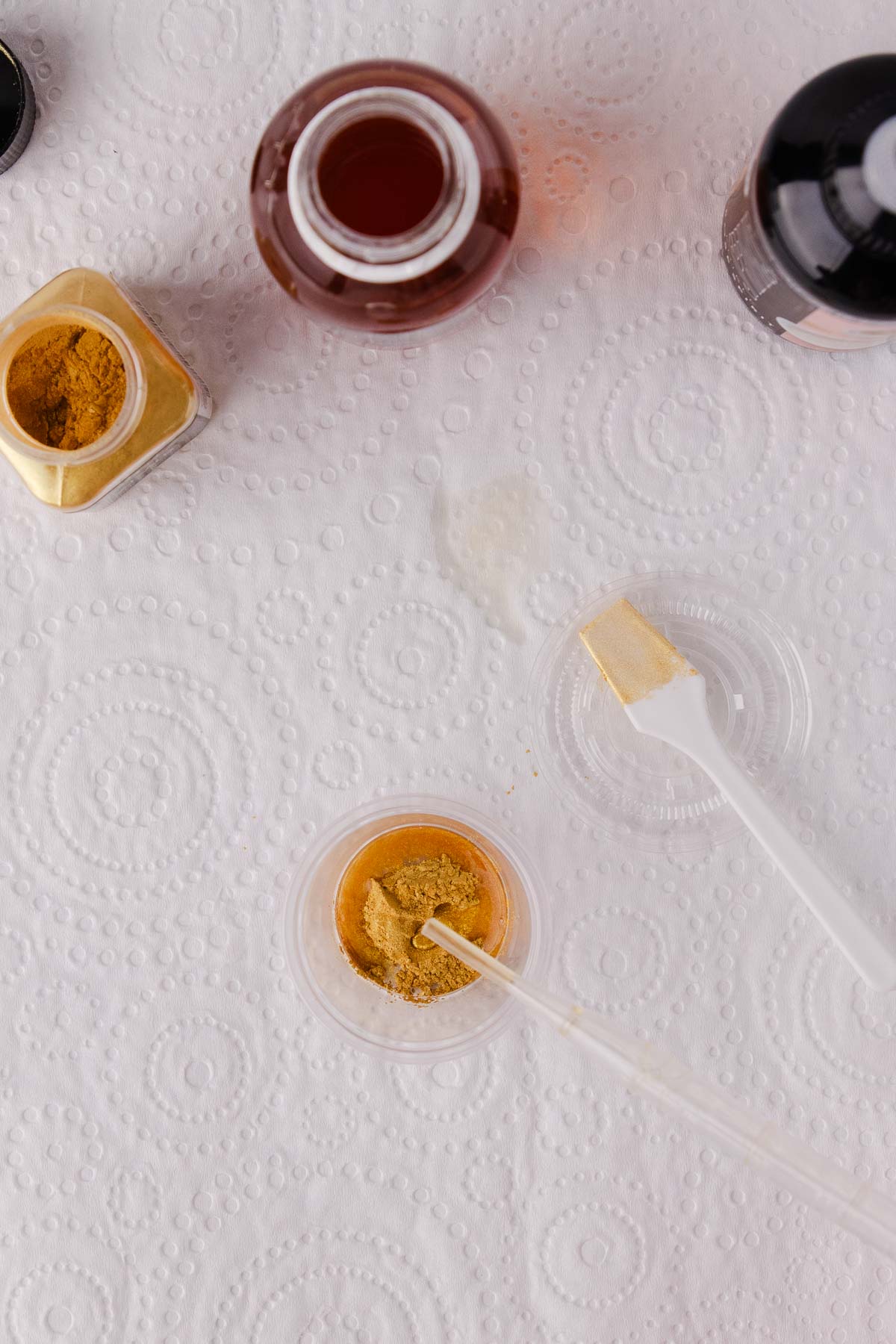 A small dropper being used to add drops of edible glaze to the gold powder in the small plastic container. 